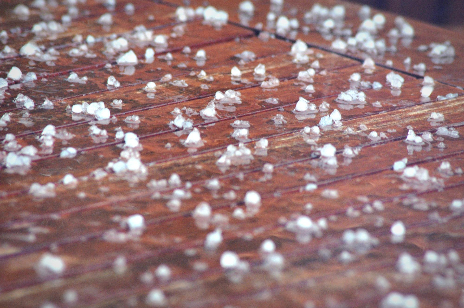 Small hailstones are scattered across a wooden deck after a hailstorm. The melting ice contrasts with the rich brown color of the wet wood.