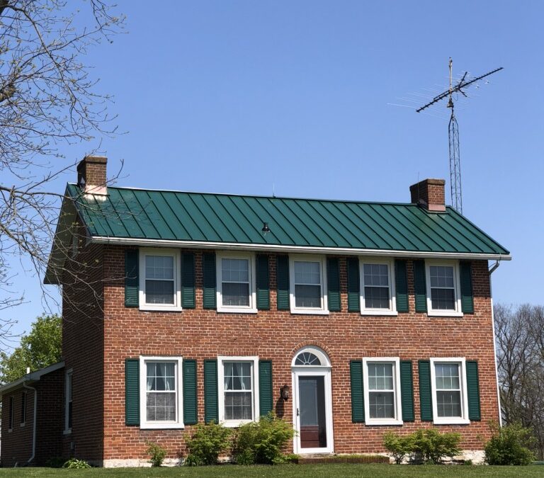 A two-story brick house with a green metal roof and green shutters stands under a clear blue sky. The lawn is well-kept and a tall TV antenna is visible beside the house.