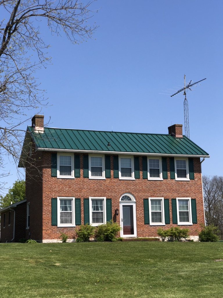 A two-story brick house with a green metal roof and green shutters stands under a clear blue sky. The lawn is well-kept and a tall TV antenna is visible beside the house.